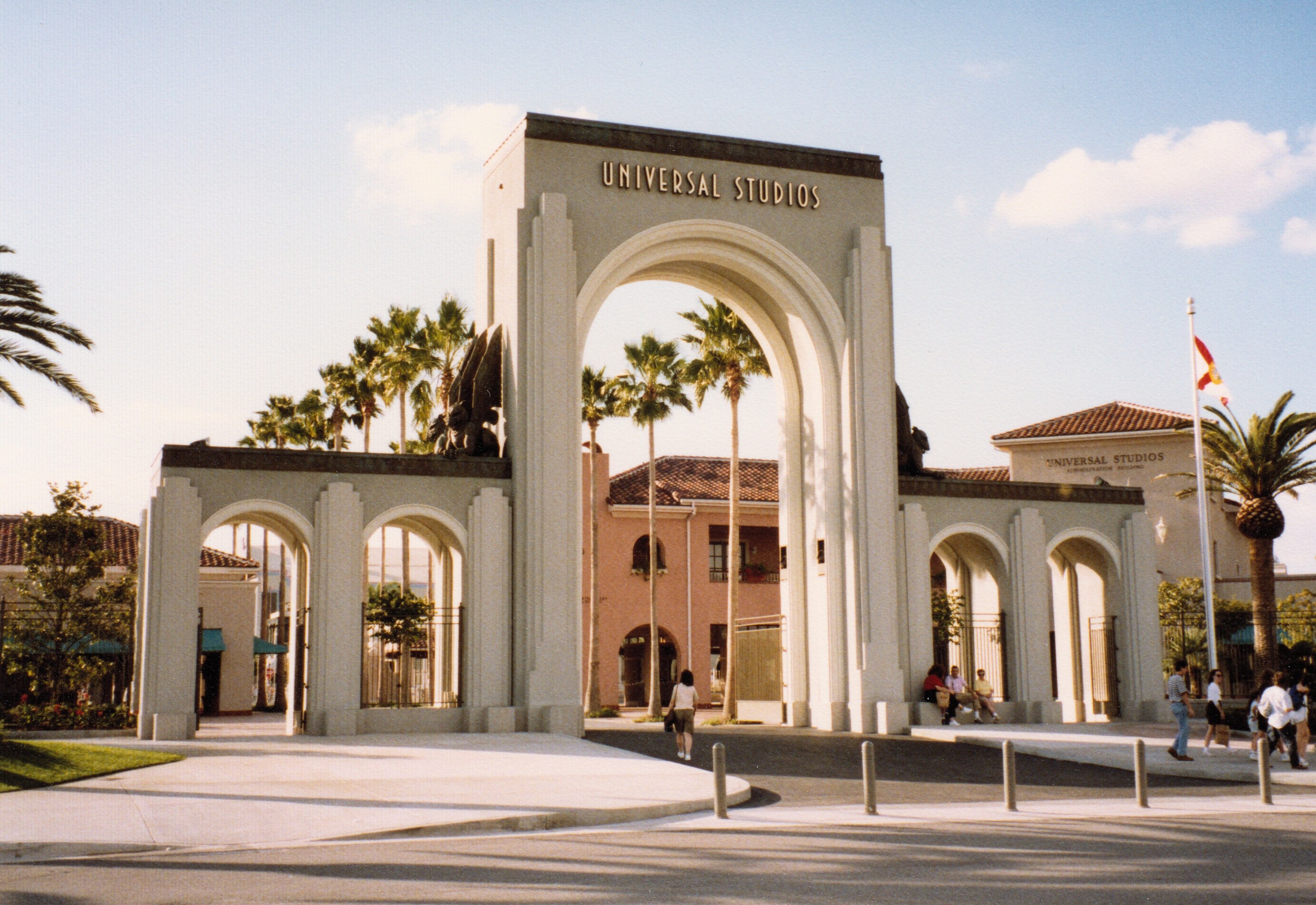 Eingang zum Universal Studios Hollywood mit einem Tor, einer Statue, Menschen, Bäumen, Gebäuden, einer Fahnenstange mit einer Fahne und einem bewölkten Himmel.