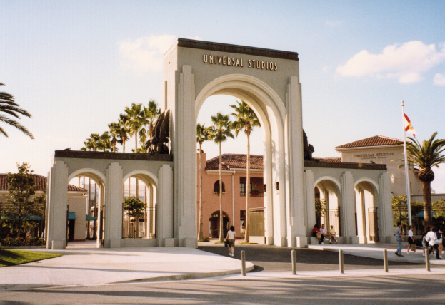 Eingang zum Universal Studios Hollywood mit einem Tor, einer Statue, Menschen, Bäumen, Gebäuden, einer Fahnenstange mit einer Fahne und einem bewölkten Himmel.