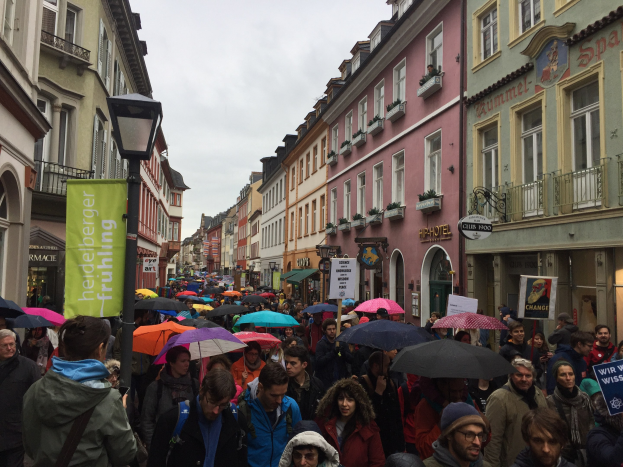 Eine große Gruppe von Menschen geht mit Schirmen die Straße entlang, einige tragen Taschen und halten Schilder, während sie an einer Klimademonstration in Deutschland teilnehmen.