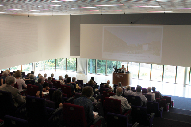 Ein Seminarraum in einer Universität mit vielen auf einen Präsentator an einem Tisch konzentrierten Zuhörern, einer auf einem Bildschirm angezeigten Präsentation und großen Fenstern mit Bäumen und Gebäuden im Hintergrund.