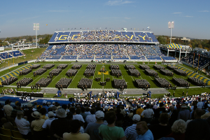 Eine große Menschenmenge in einem Stadion bei einem Fußballspiel, einige stehen, andere sitzen, auf einem grünen Feld unter einem klaren blauen Himmel, umgeben von Laternenmasten, Bäumen und Gebäuden.