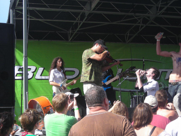 Eine Gruppe von Musikern, die verschiedene Instrumente spielen, auf einer Bühne mit einem großen schwarzen Lautsprecher links daneben, während das Publikum davor steht und die Vorstellung genießt, mit einem grünen Banner im Hintergrund.