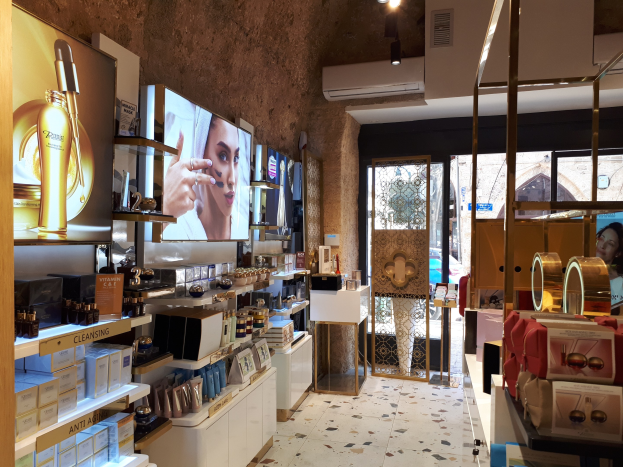 Innenansicht eines Kosmetikladens mit verschiedenen Produkten auf Regalen, Wandbildschirmen, Deckenbeleuchtung, einer Klimaanlage und Glastüren, die eine Person, ein Auto und ein Gebäude draußen zeigen.