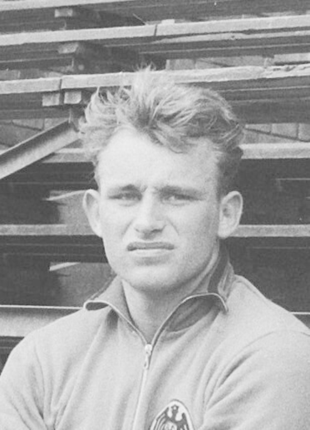 Schwarzes und weißes Foto eines Mannes in einer Jacke, der vor gestapelten Holzplanken steht, der angeblich ein legendärer deutscher Fußballspieler ist, der im Alter von 89 Jahren starb.