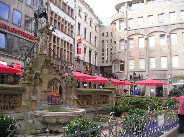 Eine Stadtansicht mit Gebäuden, Zelten und Menschen im Hintergrund und einer Skulptur, einem Teich, Pflanzen und einem Geländer im Vordergrund.