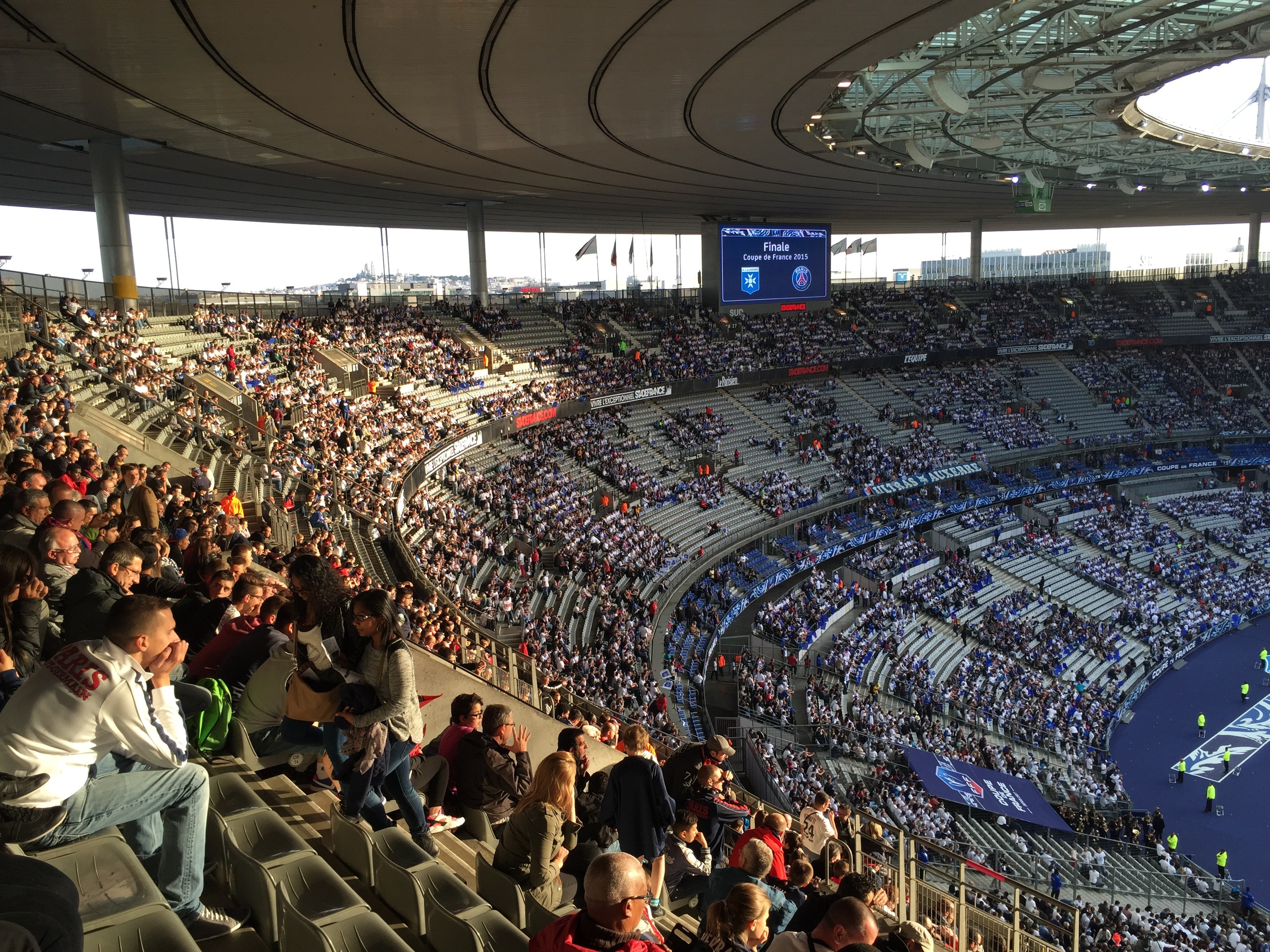 Große Menschenmenge in einem Stadion bei einem Fußballspiel mit einer Bühne, Fahnen, Stangen und einem Bildschirm im Hintergrund, wahrscheinlich die Allianz Arena in München, Deutschland.
