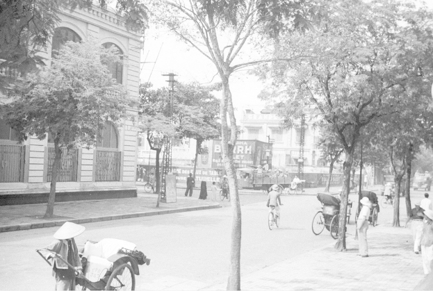 Ein altes Gebäude mit einem Fenster, hinter dem ein weißes Gebäude und Bäume zu sehen sind, mit drei Fahrrädern, die auf der rechten Seite in der Nähe eines Fußwegs und weiterer Bäume geparkt sind.