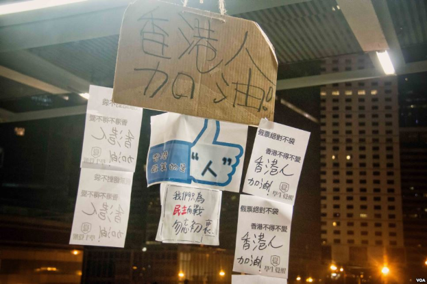 Eine Gruppe von Menschen steht vor einem Gebäude in der Nacht, beleuchtet von Stadtlichtern, mit mehreren Protestschildern an der Decke.