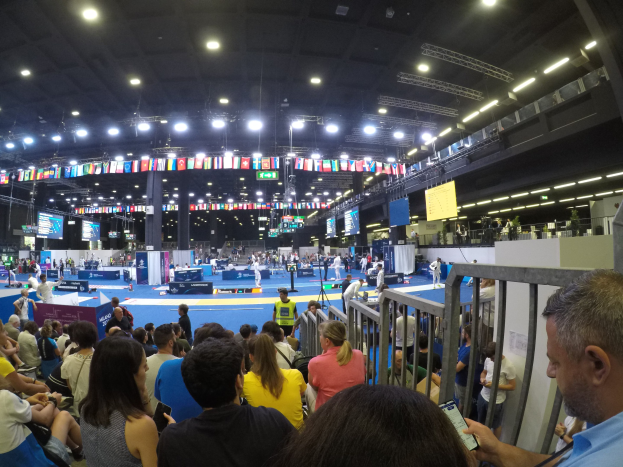 Eine große Menschenmenge sitzt und steht in einer Indoor-Sportarena mit Bildschirmen, Fahnen und Deckenbeleuchtung im Hintergrund.
