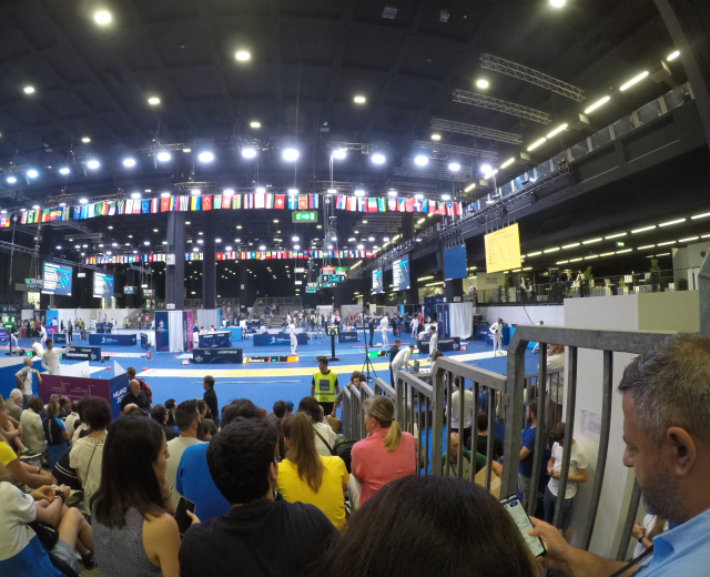 Eine große Menschenmenge sitzt und steht in einer Indoor-Sportarena mit Bildschirmen, Fahnen und Deckenbeleuchtung im Hintergrund.