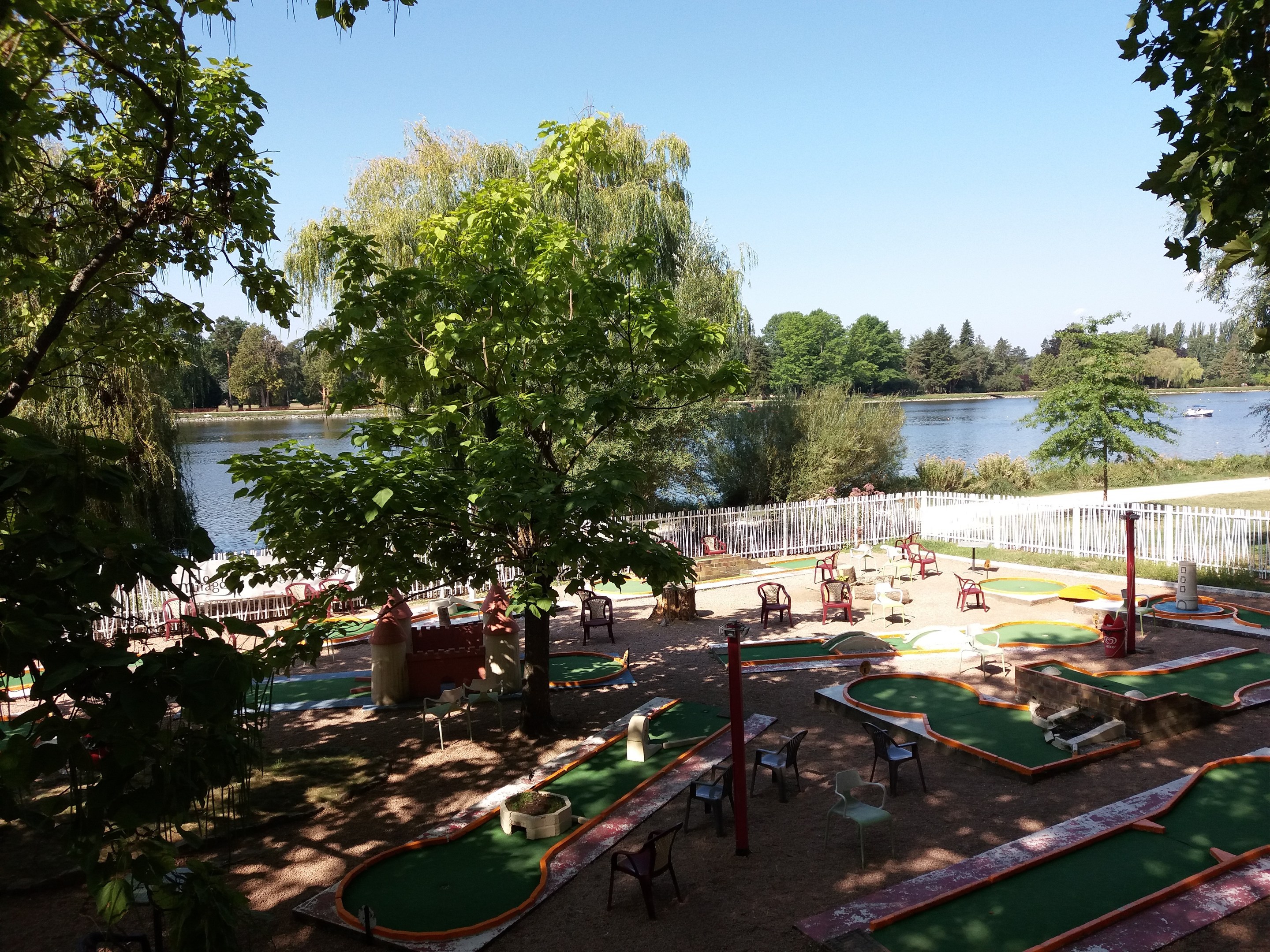 Minigolfanlage in einem Park mit grünem Gras, Bäumen, einem Zaun, verstreuten Stühlen und Tischen, einem Gewässer mit einem Boot im Hintergrund und sichtbarem Himmel.