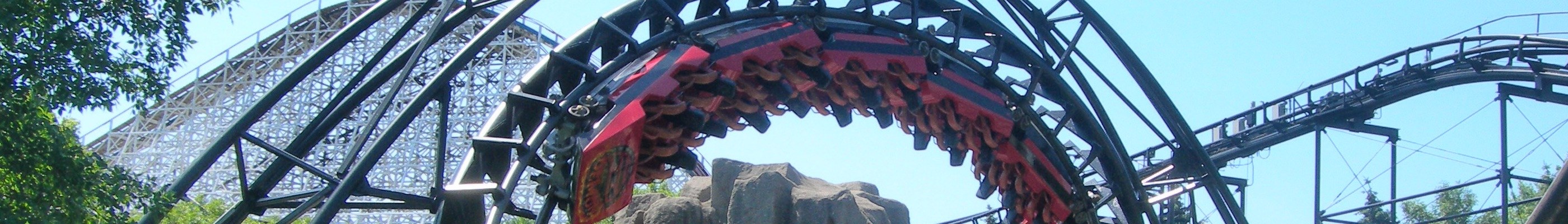 Eine Achterbahnschiene schlängelt sich durch einen von Bäumen und Felsen umgebenen Park, mit dem Himmel im Hintergrund.