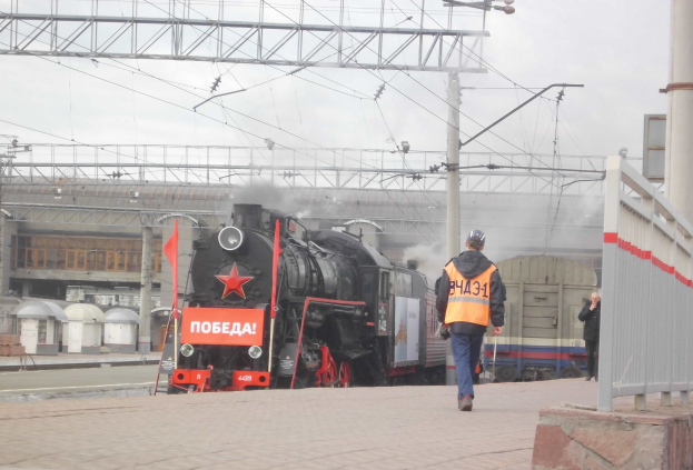 Ein Mann mit Helm und Handtasche geht auf einem Bahngleis neben einem Zug, mit einer Wand, einer Säule, Strommasten, Drähten, einem Gebäude und dem Himmel im Hintergrund.