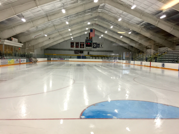 Großes Hallen-Eisstadion mit Deckenbeleuchtung, Werbetafeln, Treppen mit Geländern, einer Wand im Hintergrund und einigen Menschen, was auf eine Sportanlage hinweist, die likely für Hockey verwendet wird.