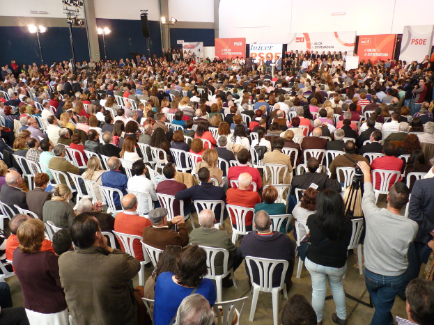 Ein volles Stadion mit vielen sitzenden und stehenden Menschen, eine Bühne mit zwei Rednern auf einem Podium und zahlreiche Banner und Lichter.