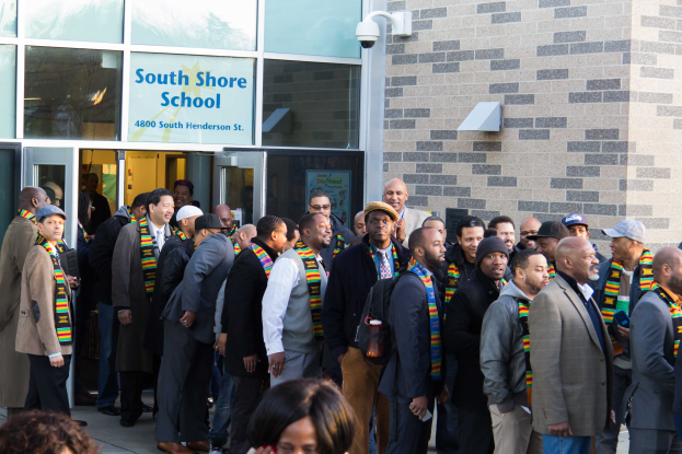 Eine Gruppe von Menschen, einige mit Mützen und Schals, steht vor dem beleuchteten South Shore School-Gebäude mit Glaswänden und Türen, versammelt zum Feiern mit einer Person, die ein Mobiltelefon und eine Texttafel hält.