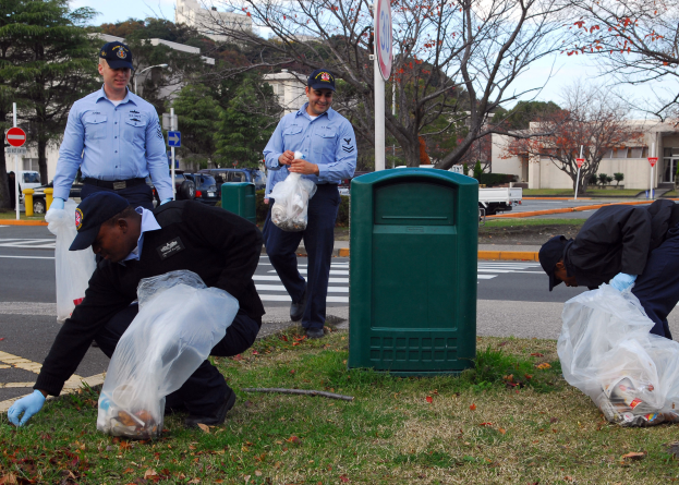 Drei Polizeibeamte in Mützen und Handschuhen sammeln Müll an einer Straßenböschung mit Bäumen, Gebäuden, Schildern, Fahrzeugen und einem Mülleimer im Hintergrund, an einem bewölkten Tag mit Gras und trockenen Blättern auf dem Boden.