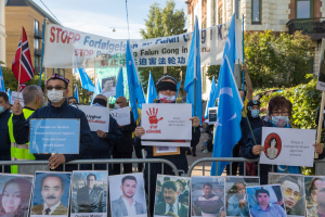Eine Gruppe von Menschen mit Masken und Fahnen und Schildern steht vor einem Zaun mit Bildern und Text, protestiert gegen die t"urkische Regierung in Istanbul, mit Geb"uden, B"umen, Pflanzen und einem Laternenpfahl im Hintergrund.