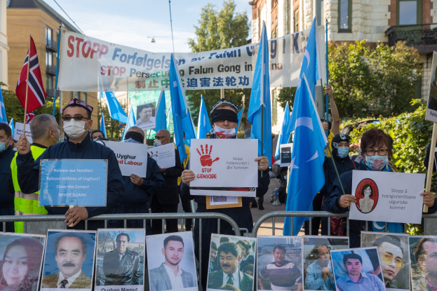 Eine Gruppe von Menschen mit Masken und Fahnen und Schildern steht vor einem Zaun mit Bildern und Text, protestiert gegen die t"urkische Regierung in Istanbul, mit Geb"uden, B"umen, Pflanzen und einem Laternenpfahl im Hintergrund.