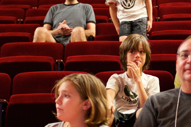Eine Gruppe von Menschen in einem Kinosaal mit roten Stühlen, mit einem Jungen in einem weißen T-Shirt und schwarzen Hosen in der Mitte.
