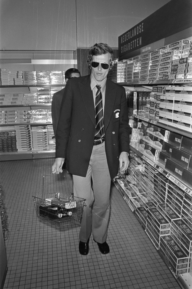 Ein Mann im Anzug und Sonnenbrille geht durch einen Laden und hält einen Einkaufswagen und eine Zigarette, umgeben von Zigarettenregalen.