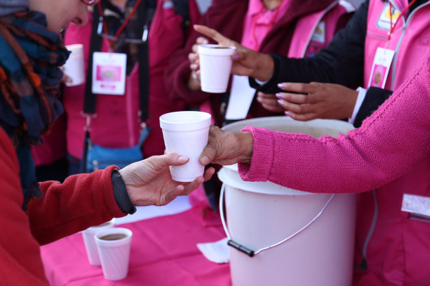 Eine Gruppe von Menschen um einen Tisch mit einem pinken Eimer herumstehend, jeder hält einen Kaffeebecher in der Hand, mit mehreren Bechern auf dem Tisch, was auf die Teilnahme an einer Brustkrebs-Awareness-Veranstaltung hindeutet.