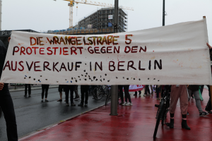 Eine Gruppe von Menschen steht auf einer Straße und hölt ein Banner mit der Aufschrift "Die Wrangellstraße 5 protestiert gegen den Ausverkauf in Berlin". Im Hintergrund sind Fahrráder, Masten, Gebáude und ein Kran zu sehen, öber ihnen der Himmel.