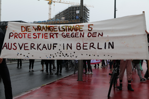 Eine Gruppe von Menschen steht auf einer Straße und hölt ein Banner mit der Aufschrift "Die Wrangellstraße 5 protestiert gegen den Ausverkauf in Berlin". Im Hintergrund sind Fahrráder, Masten, Gebáude und ein Kran zu sehen, öber ihnen der Himmel.