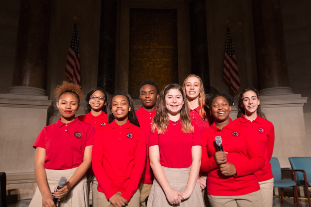 Gruppe junger Frauen in roten Shirts, die strahlend Mikrofone halten und Teil eines nationalen Jugendchors sind, mit Stühlen, Säulen, Fahnen und einer Wand im Hintergrund.