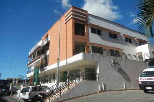 Ambulanz vor einem Gebäude mit Fenstern, Geländern, Treppen und einer Treppe geparkt, mit Fahrzeugen auf der Straße, einem Baum auf der rechten Seite, Strommasten mit Drähten und einem bewölkten Himmel im Hintergrund.