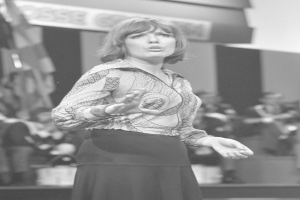 Eine Frau in einem Schwarz-Weiß-Foto steht auf einer Bühne umgeben von einer Gruppe von Menschen, trägt ein weißes Hemd und einen schwarzen Rock und hat ihr Haar zu einem Dutt gebunden und einen ernsten Gesichtsausdruck.