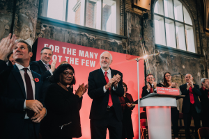 Eine Gruppe von Menschen steht vor einem Rednerpult und klatscht begeistert, mit einer Tafel mit Text auf dem Rednerpult und einem Banner mit Text an der Wand dahinter, sowie Stühlen, die im Raum verteilt sind.