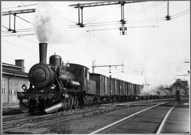 Schwarzes und weißes Foto eines Dampflokomotivs auf Schienen mit Rauch aus seinem Motor, ein Gebäude auf der linken Seite und Strommasten mit Drähten im Hintergrund.