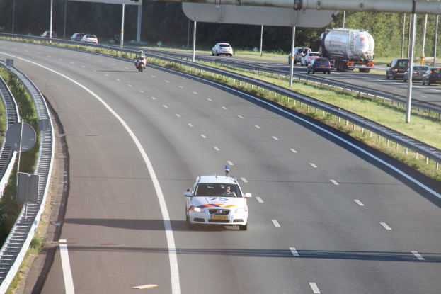 Ein Auto auf einer Autobahn mit einem stationären Polizeiwagen am Straßenrand, andere Fahrzeuge, Leitplanken, Pfosten, Schilder und Bäume im Hintergrund.
