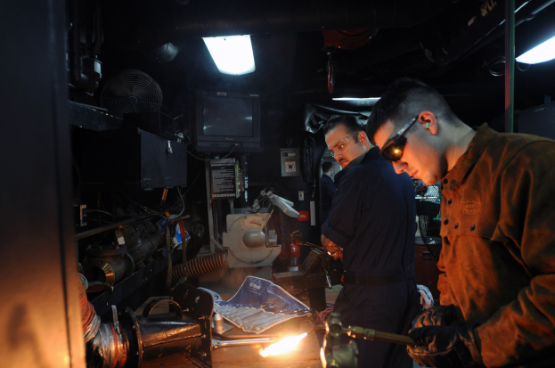 Zwei Männer mit Brille arbeiten an einem Metallteil in einer Werkstatt, einer verwendet ein Schweißgerät; Werkzeuge und Ausrüstung liegen auf dem Tisch, mit einem Fernseher und Lampen im Hintergrund.