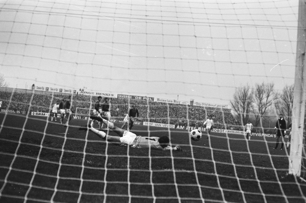 Schwarzes und weißes Foto eines Fußballspielers, der einen Ball vor einem Tor abwehrt, mit einer Menge, einem Banner, Bäumen und einem bewölkten Himmel im Hintergrund.