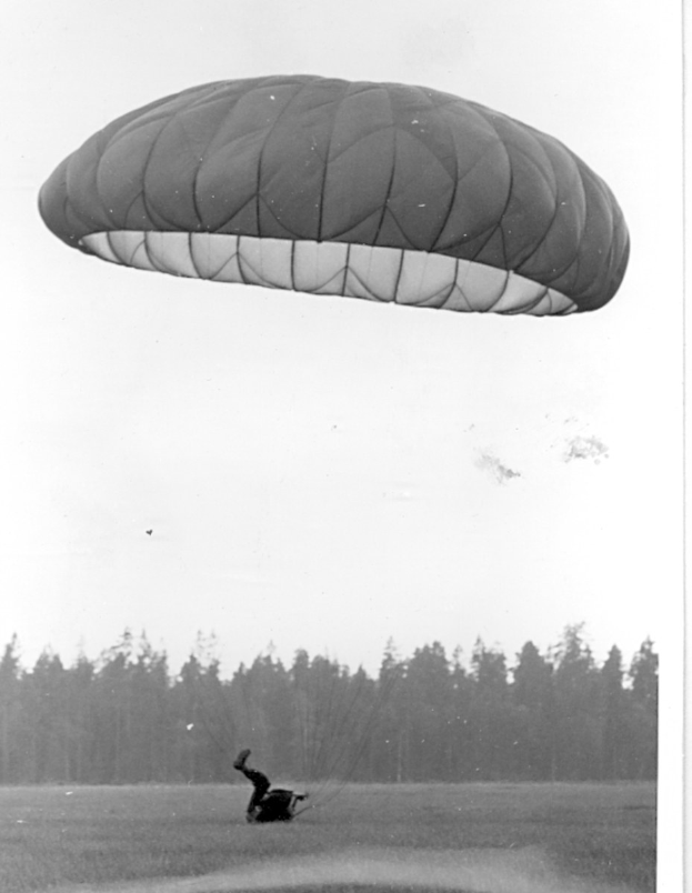 Ein Schwarz-Weiß-Foto einer Person, die mit einem Fallschirm paragleitet, umgeben von Bäumen und einem klaren Himmel, mit Text unten.