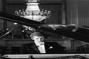 Ein Mann in einem Smoking, der vor einem Kronleuchter auf einem Klavier spielt, umgeben von Pflanzen und einer Wand, auf einem Schwarz-Weiß-Bild.