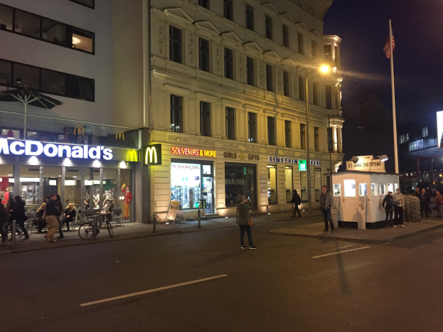 Eine Gruppe von Menschen geht nachts eine Straße in Berlin, Deutschland, entlang mit Gebäuden, Laternen, einer Fahne, Schaufenstern, Fahrrädern und dem Himmel im Hintergrund.