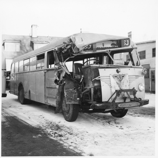 Schwarzes und weißes Foto eines schwerbeschädigten Busses mit eingedellter Front und zersplitterten Fenstern am Straßenrand, mit Gebäuden im Hintergrund.