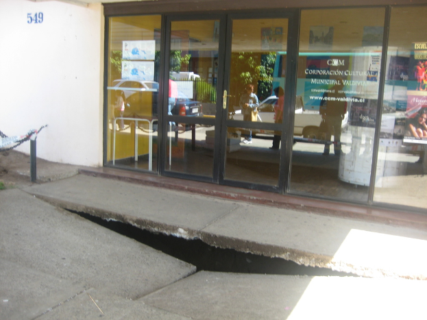 Eine zerbrochene Fläche im Vordergrund mit einer Glastür, Wand und einem Pfahl im Hintergrund, die sich bewegende Fahrzeuge und einige stehende Personen spiegeln.