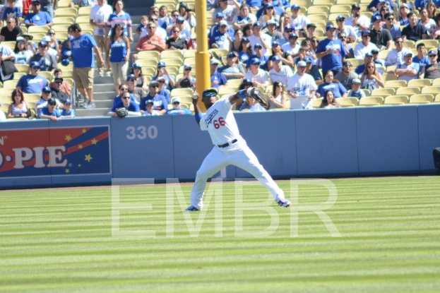 Ein Mann mit Mütze und Sonnenbrille versucht, einen Ball auf einem Rasenfeld zu werfen, umgeben von einem Publikum, das auf Stühlen sitzt und steht, wobei einige ebenfalls Mützen tragen, und ein Pfahl ist im Hintergrund sichtbar.