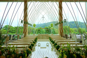 Eine Hochzeitszeremonie in einem großen Gebäude mit in einem Halbkreis angeordneten Tischen und Stühlen, geschmückt mit Blumenbouquets, mit Bäumen, Hügeln und einem klaren blauen Himmel im Hintergrund.