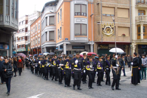 Eine Gruppe von Menschen, die mit Schirmen die Straße entlanggehen, einige halten Musikinstrumente und andere tragen Taschen, mit Gebäuden, Laternenmasten und Namensschildern im Hintergrund, unter einem sichtbaren Himmel, während eines Ehrenzuges.