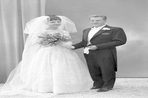 Ein Schwarz-Weiß-Foto eines Mannes und einer Frau, die Hand in Hand auf ihrer Hochzeit stehen, wobei die Frau ein weißes Kleid trägt und einen Blumenstrauß hält und der Mann strahlt, mit einem Vorhang und einer Wand im Hintergrund.