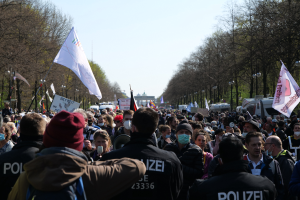 Eine große Gruppe von Menschen steht vor einer Menge von Polizeibeamten, einige tragen Mützen und Masken, während sie bei einer Demonstration in Berlin, Deutschland, mit Schildern, Fahnen, Laternenmasten, Bäumen, Fahrzeugen, einem Gebäude und dem Himmel im Hintergrund demonstrieren.