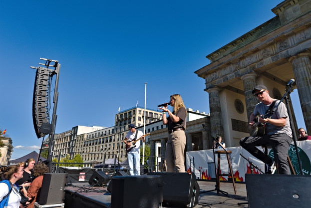 Eine Gruppe von Menschen, die auf einer Bühne Musik spielen, mit dem Brandenburger Tor im Hintergrund, begleitet von Lautsprechern und anderem Equipment unter einem klaren blauen Himmel.