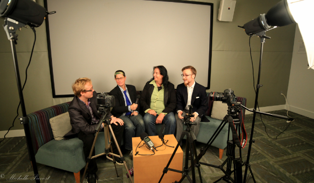 Eine Gruppe von drei Personen sitzt auf einem Sofa und gibt ein Interview, wobei eine Person in einem schwarzen Mantel und Hose links, eine Person in einem Mantel und blauer Krawatte mit einem Mikrofon in der Mitte und eine Person mit einem Lächeln rechts sitzt. Ein Mikrofonrecorder befindet sich auf dem mittigen Tisch, Kameras sind auf der rechten und linken Seite und große Scheinwerfer sind vorhanden. Ein Projektionsschirm befindet sich im Hintergrund.