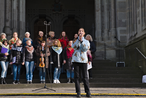 Eine Gruppe von Menschen steht vor einem Gebäude, eine Frau in der Mitte hält ein Mikrofon und singt, umgeben von Mikrofonen mit Ständern, Musikinstrumenten, Treppen mit Geländern und einer Wand mit Säulen und einer Tür auf der rechten Seite.
