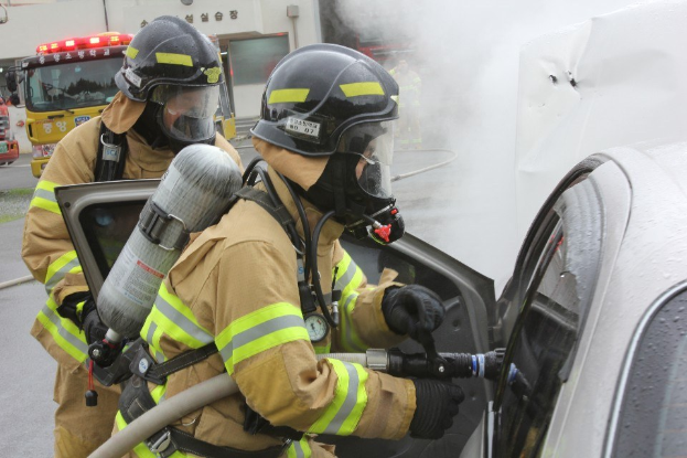 Feuerwehrleute in Schutzausrüstung verwenden einen Schlauch, um ein brennendes Auto zu löschen, mit Rauch sichtbar und Fahrzeugen und einem Gebäude im Hintergrund.